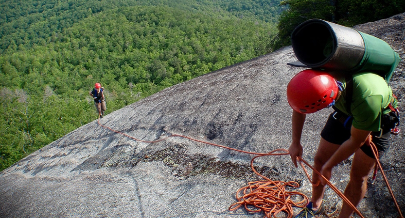 In the foreground, a person wearing safety gear is secured by ropes as they look down a rock incline at another person wearing safety gear, making their way up. On the ground below is a thick, green wooded area.